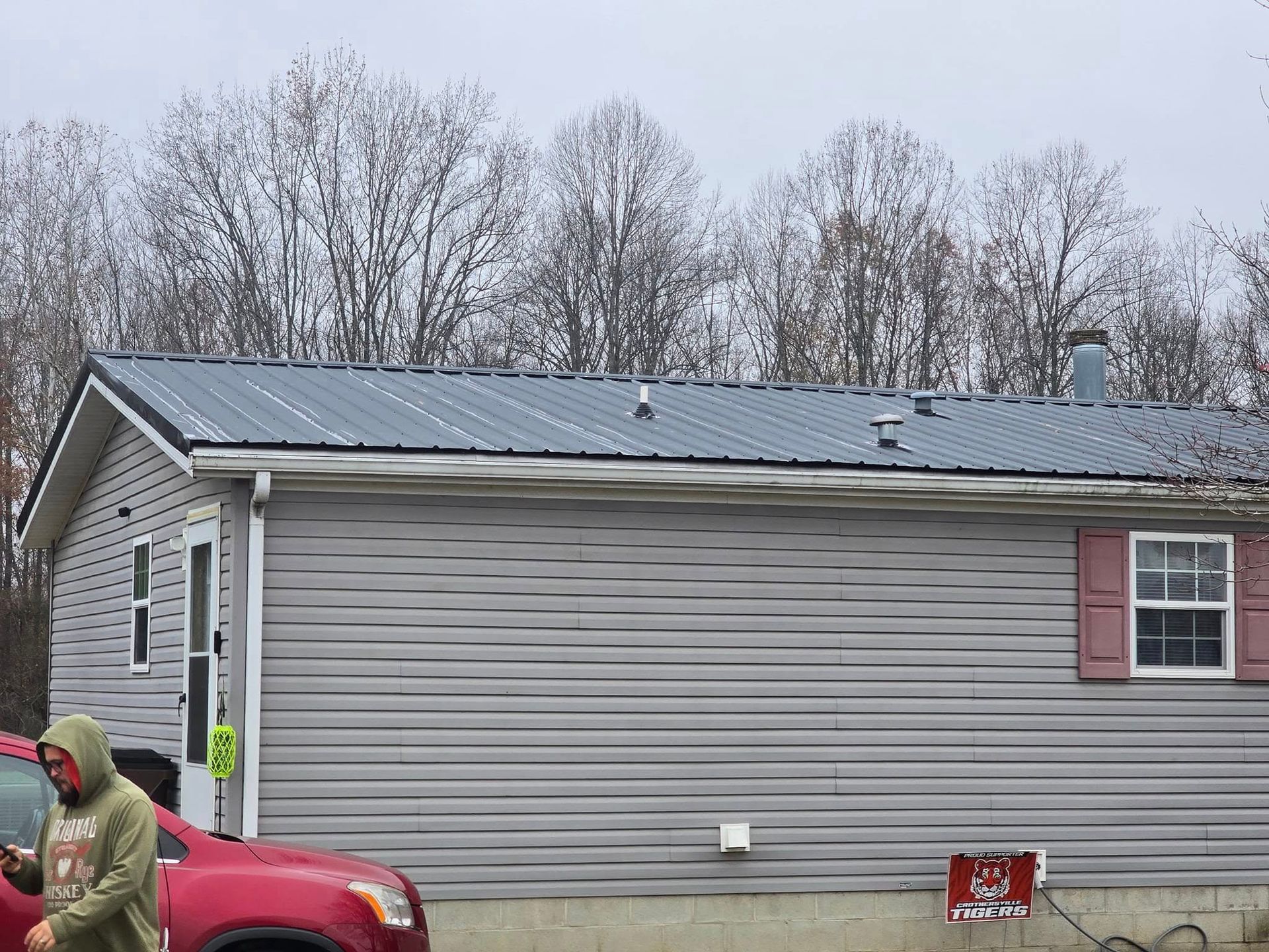 A person in a green hoodie walks past a grey vinyl-sided house with a dark metal roof and red window shutters.