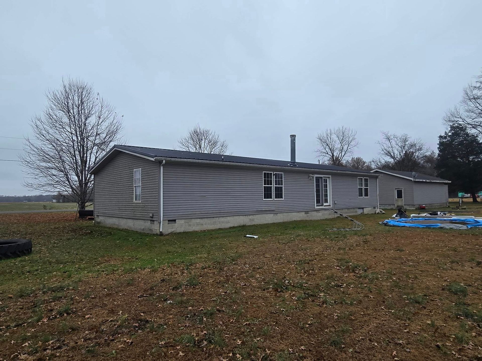 A long, grey single-story manufactured home on a rural lot under an overcast sky, with a blue tarp on the ground nearby.