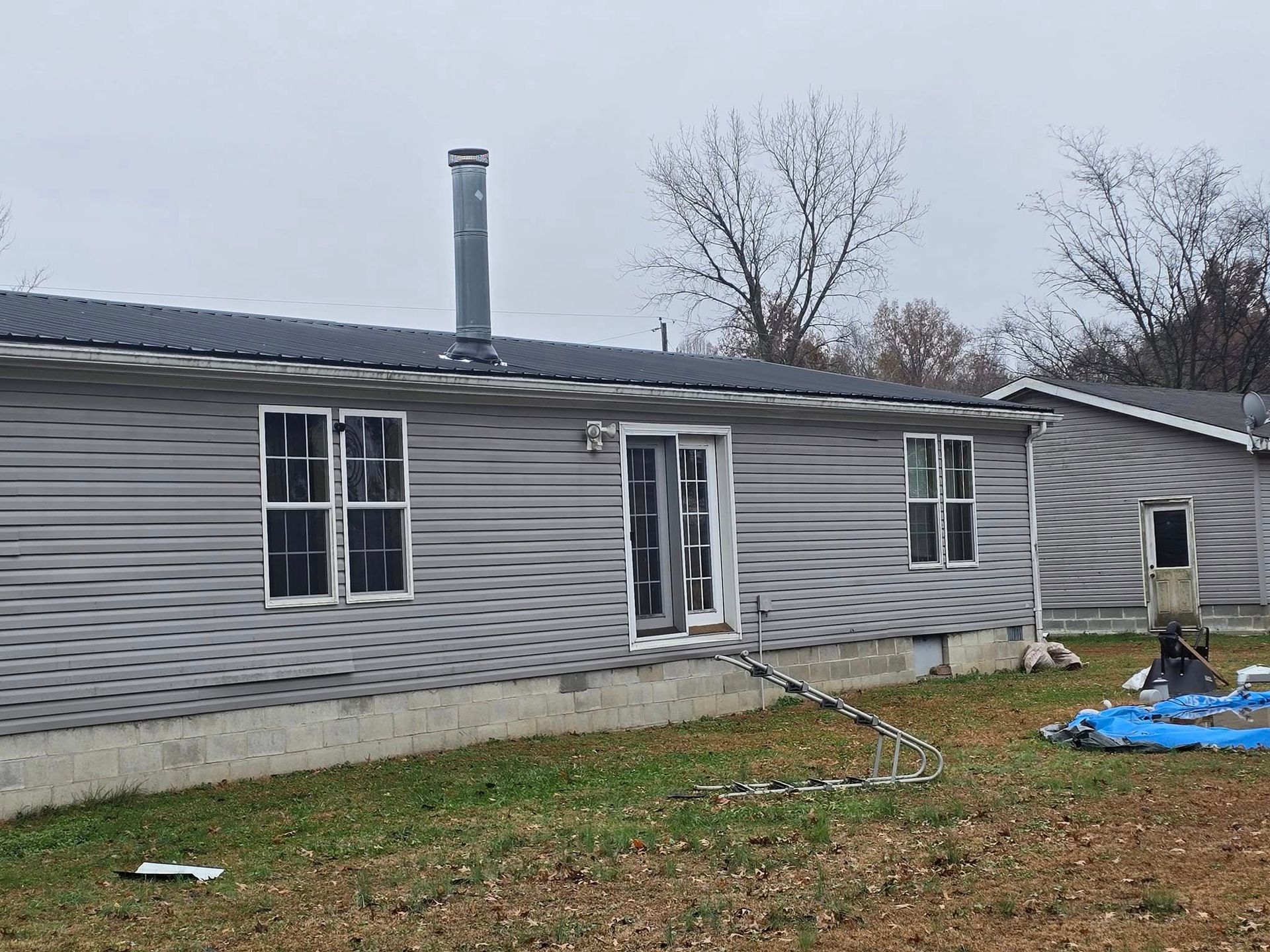 A gray, single-story mobile home with a metal roof and chimney, set against a backdrop of trees under a cloudy sky.