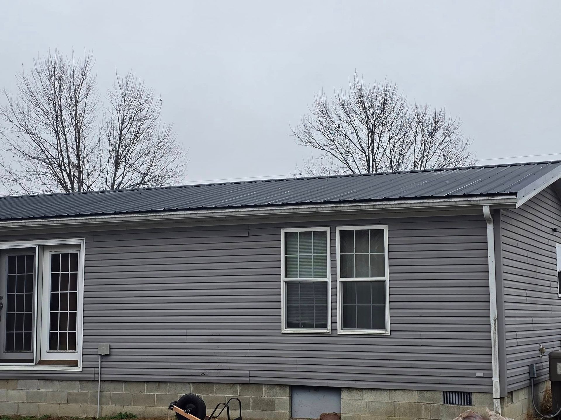 Single-story house with gray horizontal siding, a dark metal roof, white trim, and a concrete block foundation.