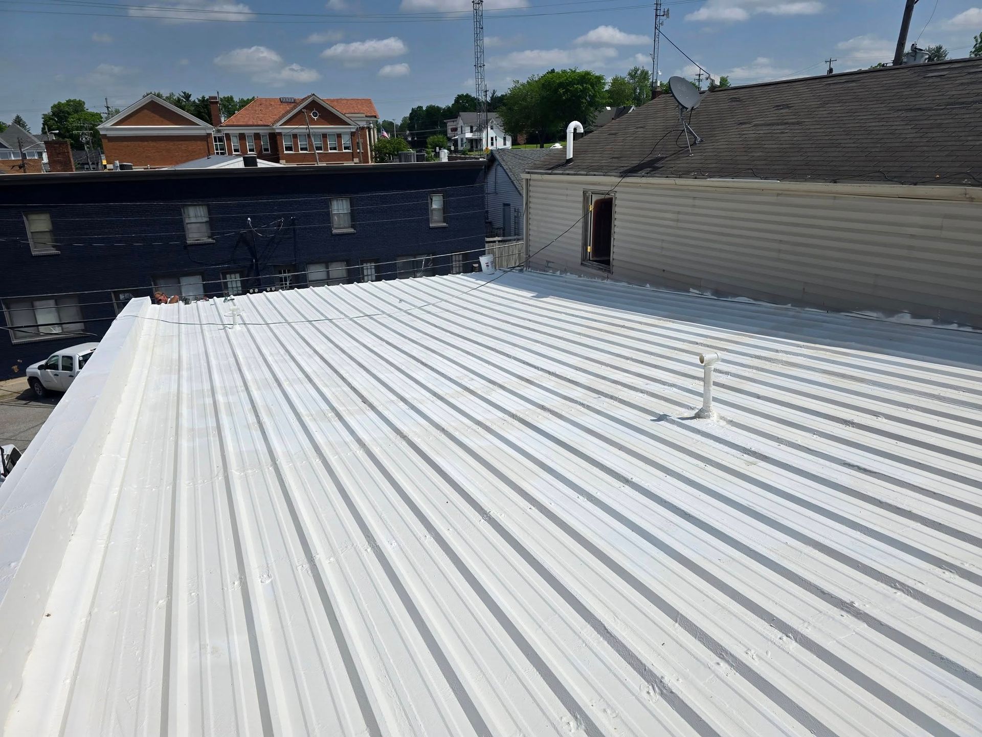A high-angle view of a white, corrugated metal roof on a sunny day, with neighboring buildings in the background.