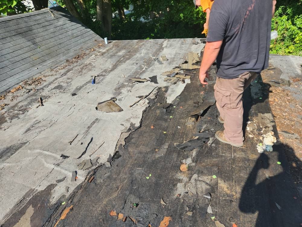 A worker stands on a partially stripped residential roof, showing exposed underlayment and debris.