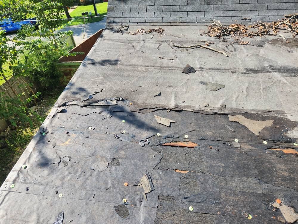 A damaged, weathered flat roof showing exposed dark underlayment and debris with a brick wall in the background.