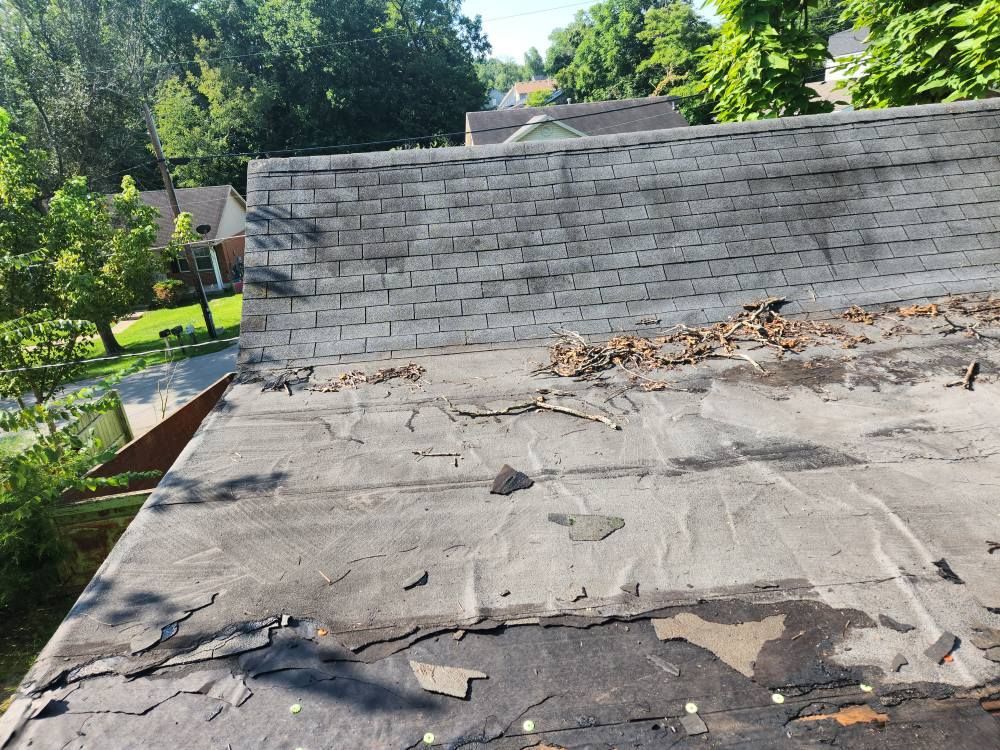 A low-slope roof section with damaged, peeling roofing material below a steep, shingled roof pitch and trees.