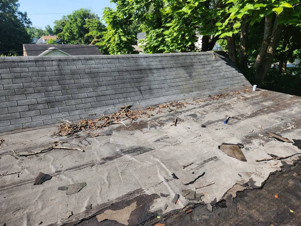 A weathered, multi-layered roof showing damaged shingles, exposed underlayment, and scattered debris near trees.