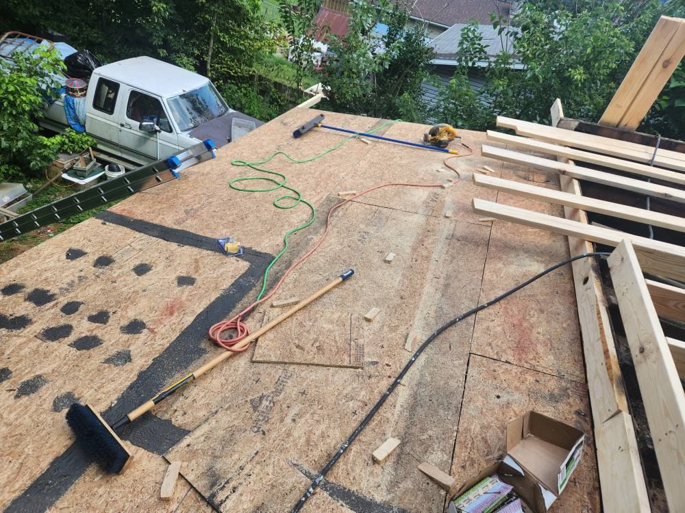 Construction scene on a plywood-covered roof with scattered tools, materials, and a white truck parked below.