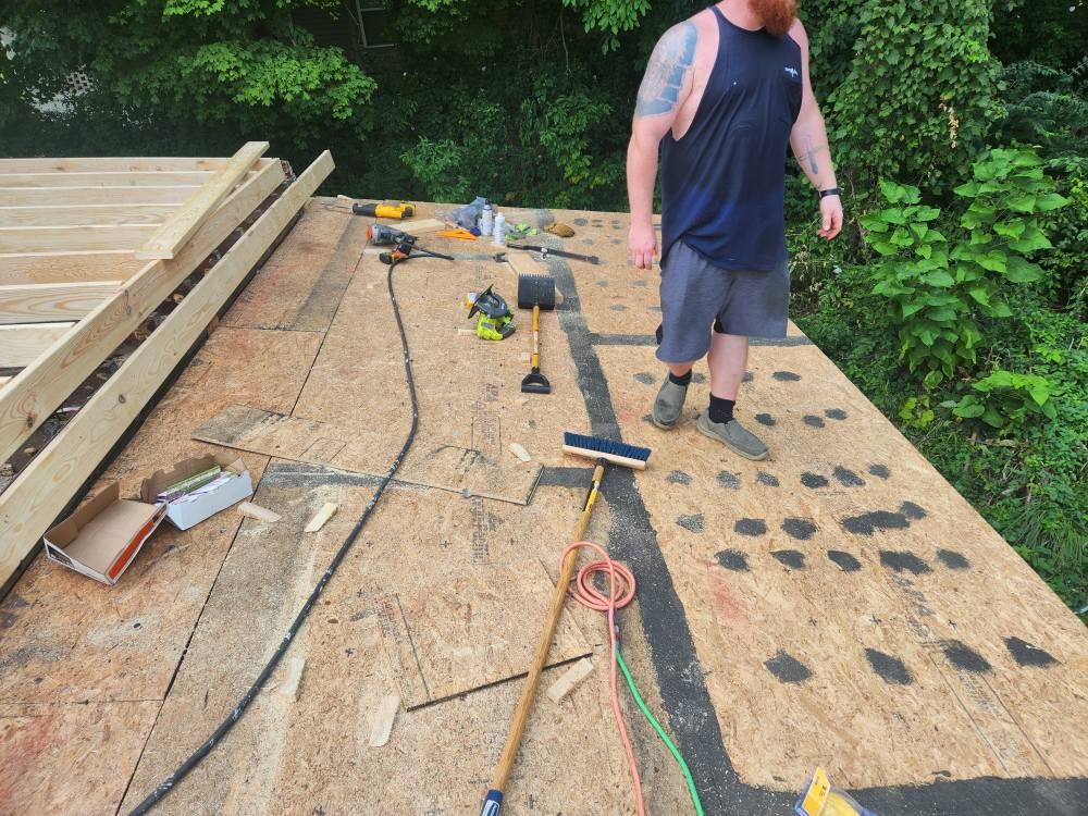 A person stands on a wooden roof deck under construction, surrounded by tools and construction materials.