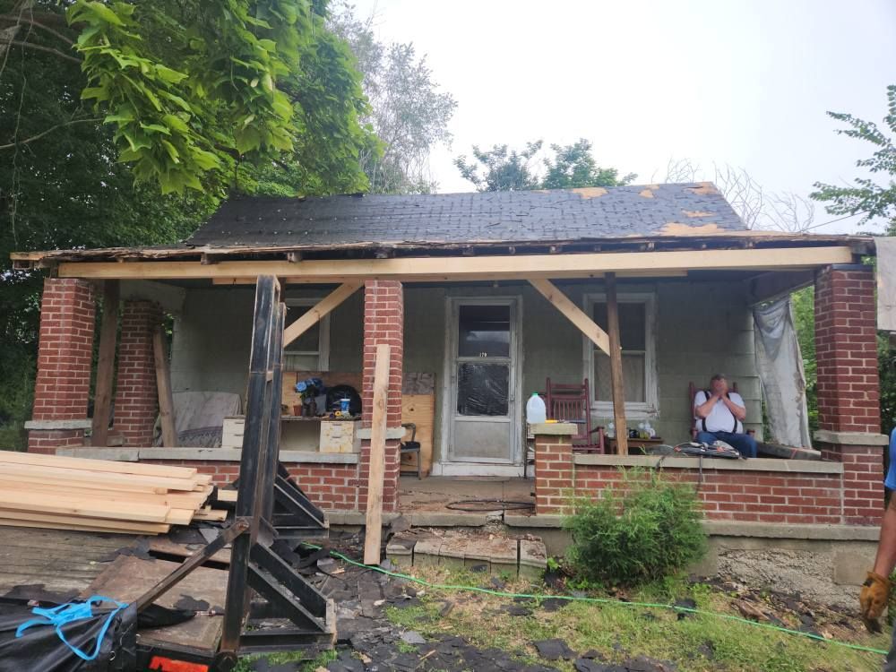 A one-story brick house undergoing front porch renovations, with construction materials and a person sitting on the porch.