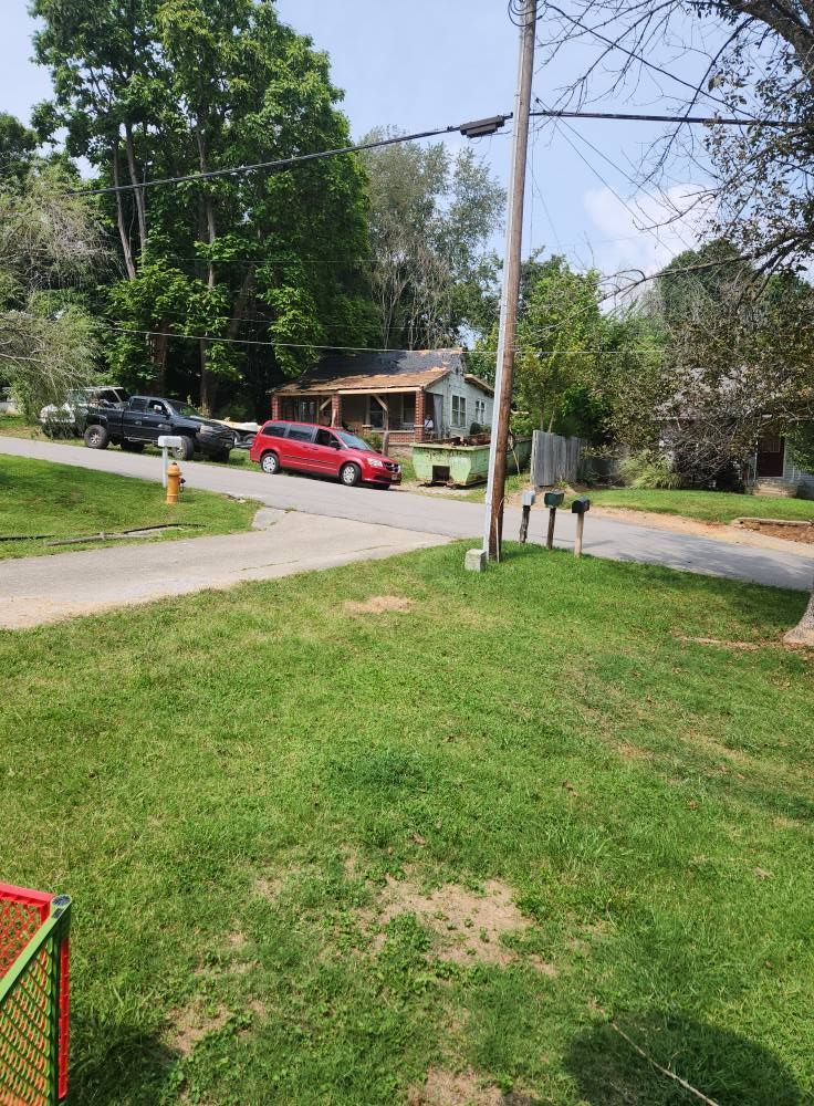 A red station wagon drives past a house and a dumpster on a quiet street with a utility pole in the foreground.