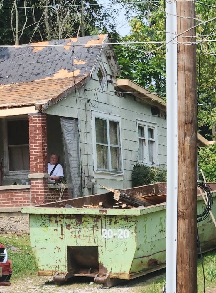 A person sits on the porch of a house undergoing renovations with a large, green construction dumpster in the yard.
