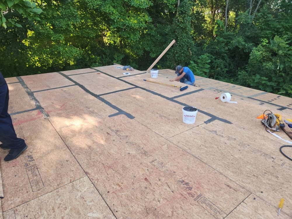 A worker kneels on a roof sheathed with plywood panels, applying tape to the seams with tools and supplies nearby.