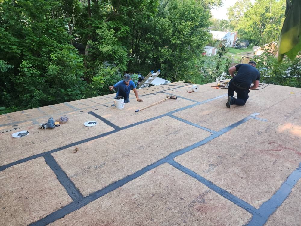 Two workers prepare a residential roof deck, applying black sealant tape between wood sheathing panels.