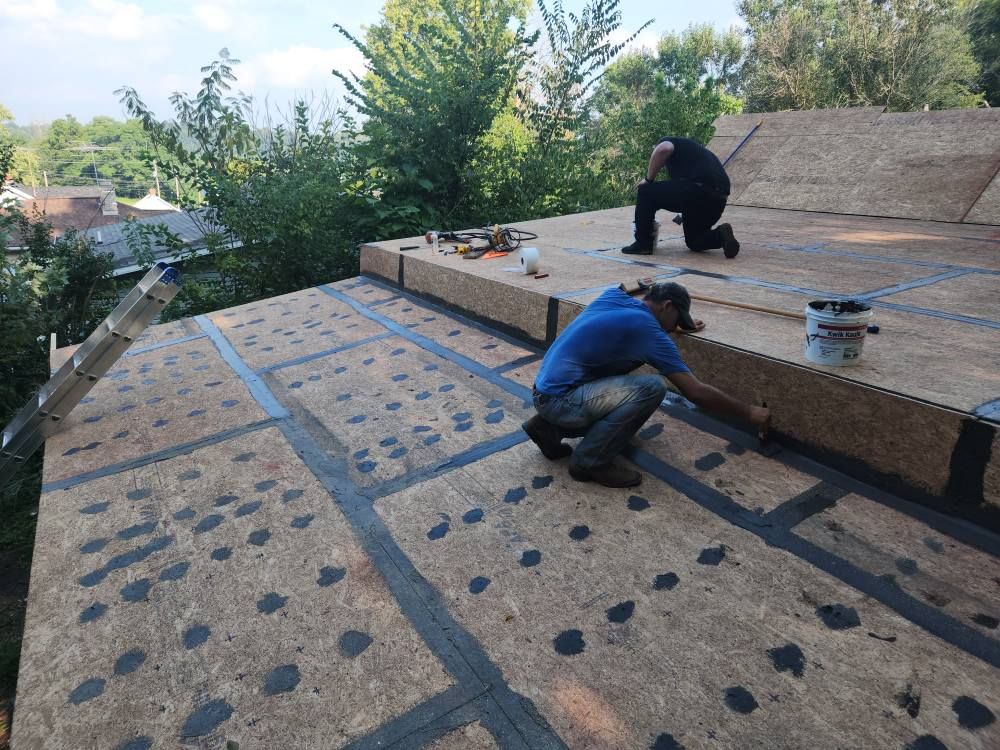 Two workers install roofing material on a plywood-decked roof, applying sealant along the seams.