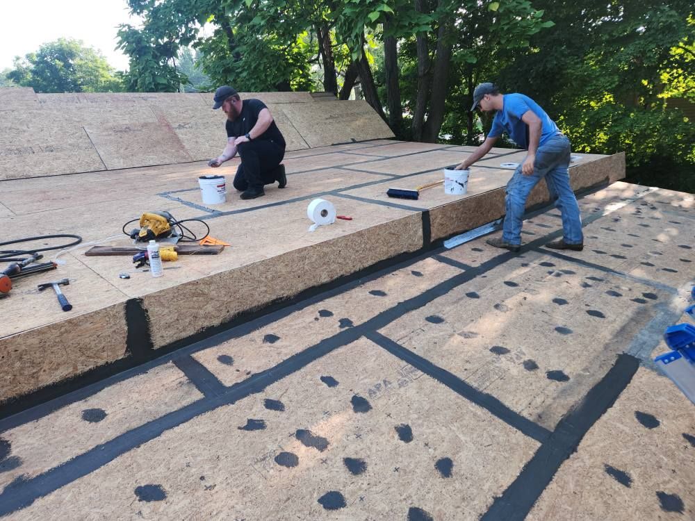 Two workers apply sealant and tape to seams on a plywood roof deck under a clear sky.
