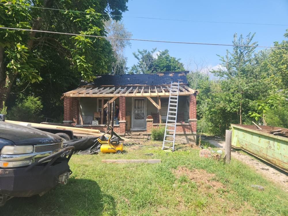 A house with a brick porch under construction, featuring exposed wooden roof rafters, a ladder, and a dump trailer nearby.