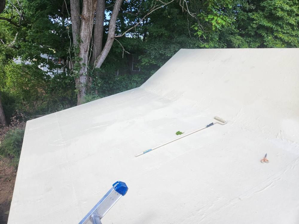 A paint roller and scissors rest on a freshly primed, light-colored wooden roof outdoors, with trees in the background.
