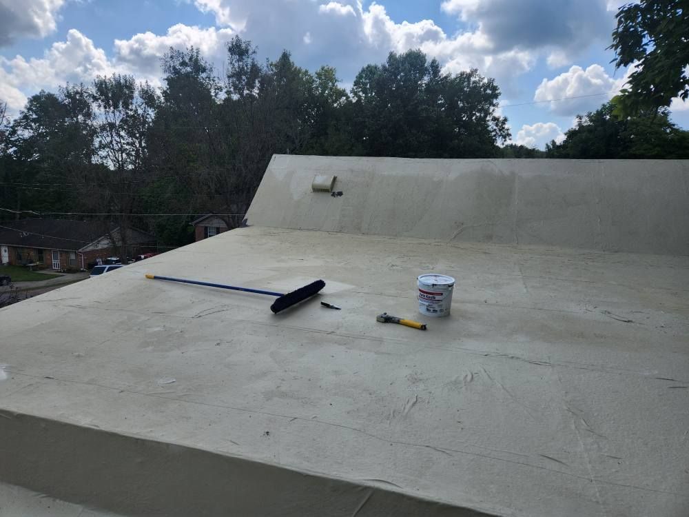 A flat rooftop covered in light-colored coating with a long-handled brush, a small paint container, and a trowel nearby.