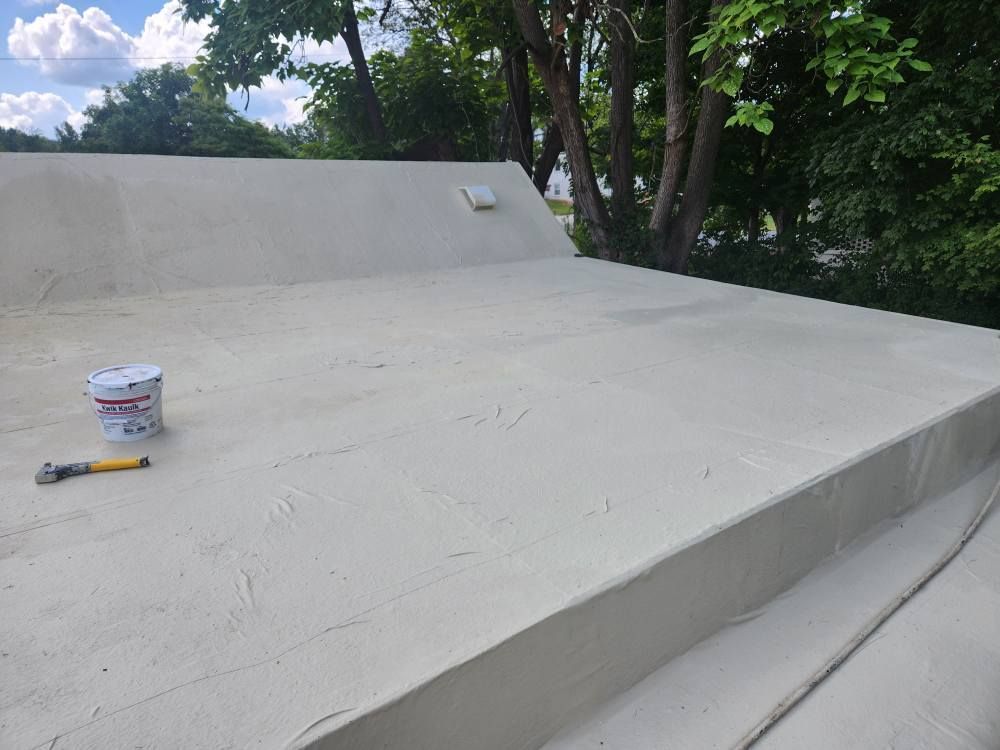 A partially coated, light-colored flat roof features a bucket and tools on the surface near trees under a blue sky.