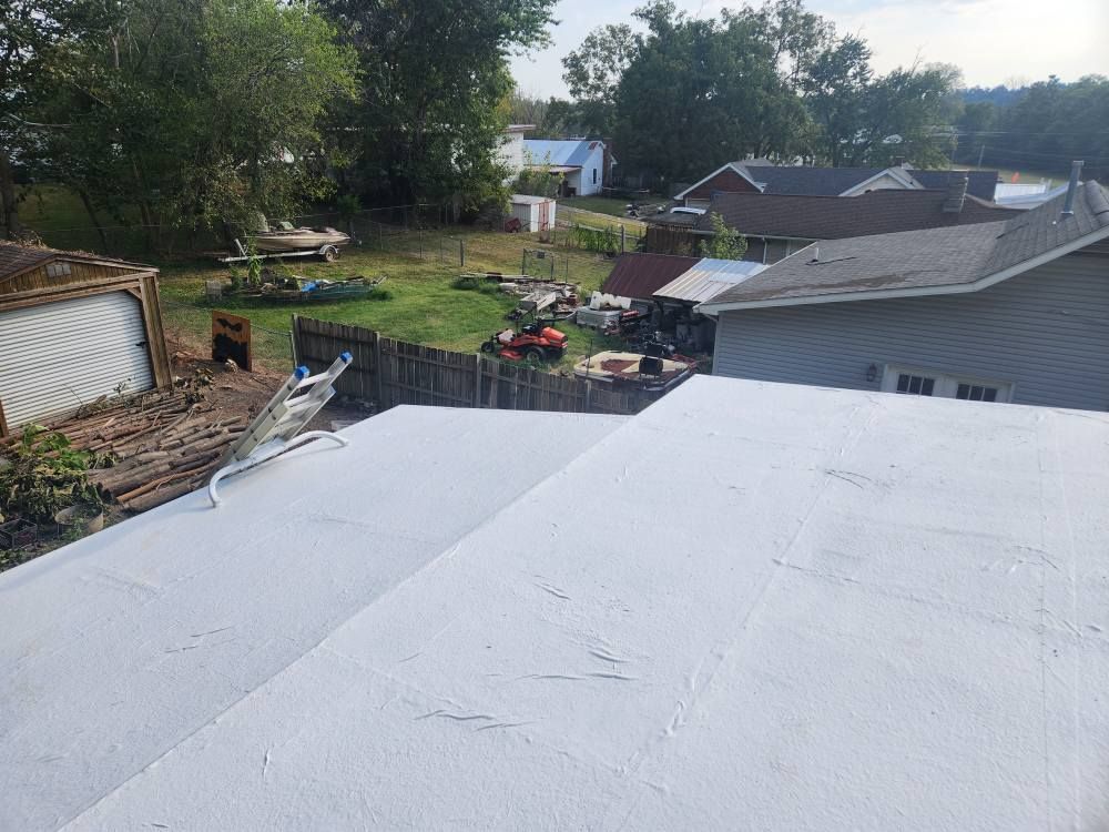 A view from a flat, white roof overlooking a backyard with trees, houses, and scattered items on a sunny day.