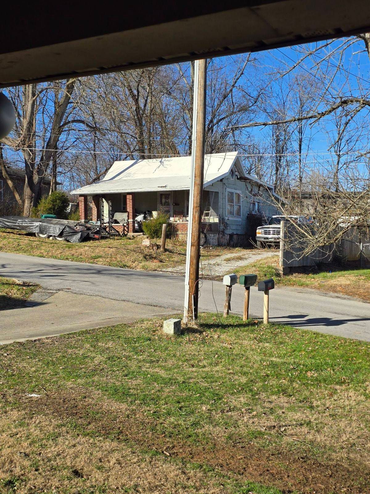 A small, weathered house with a white roof and porch sits across a road, viewed from a grassy lawn with three mailboxes.