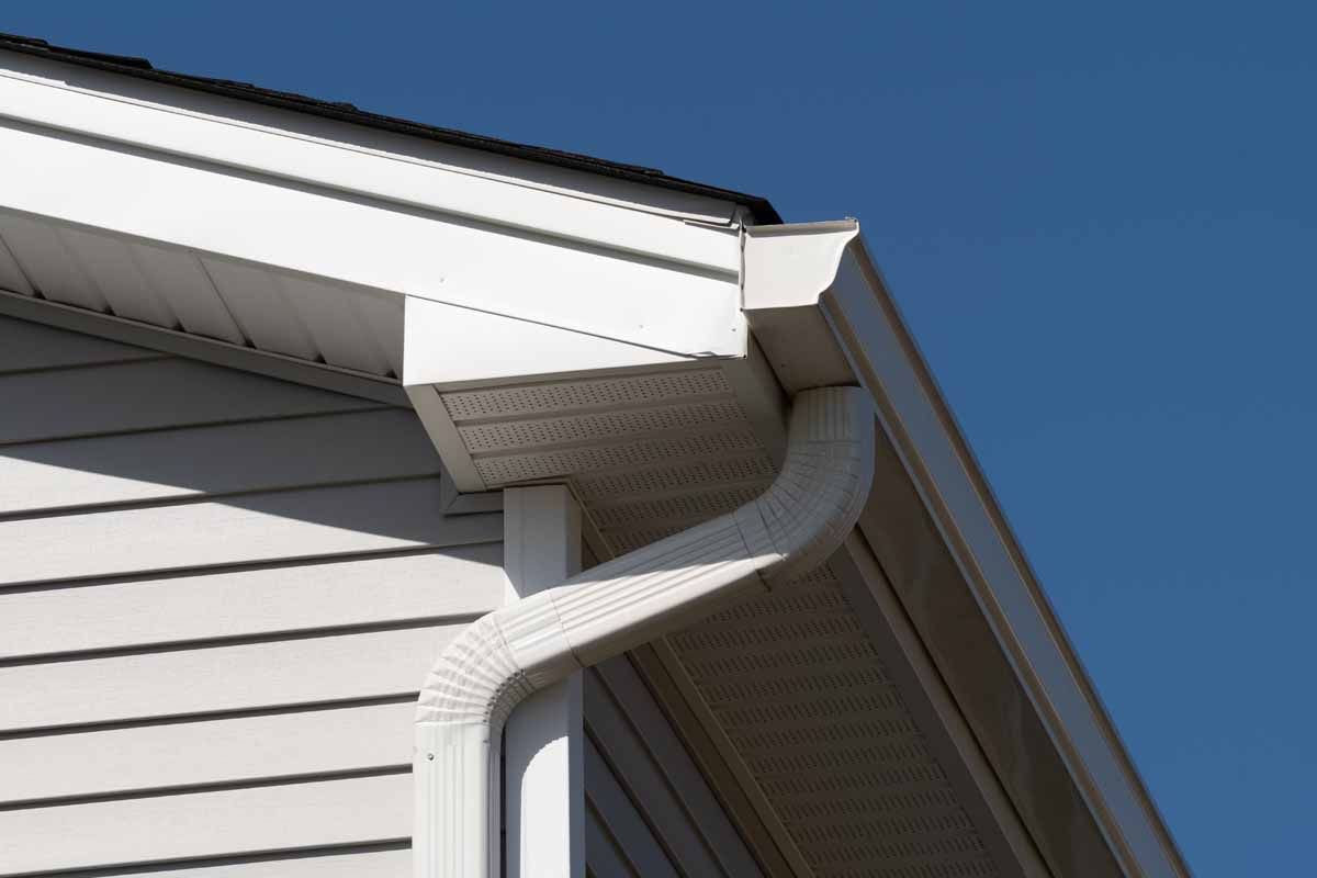 White rain gutter and downspout attached to the corner of a house with beige siding under a blue sky.