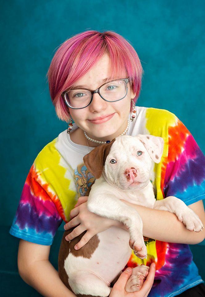 A smiling tween girl with pink hair and glasses holds her puppy to her chest.