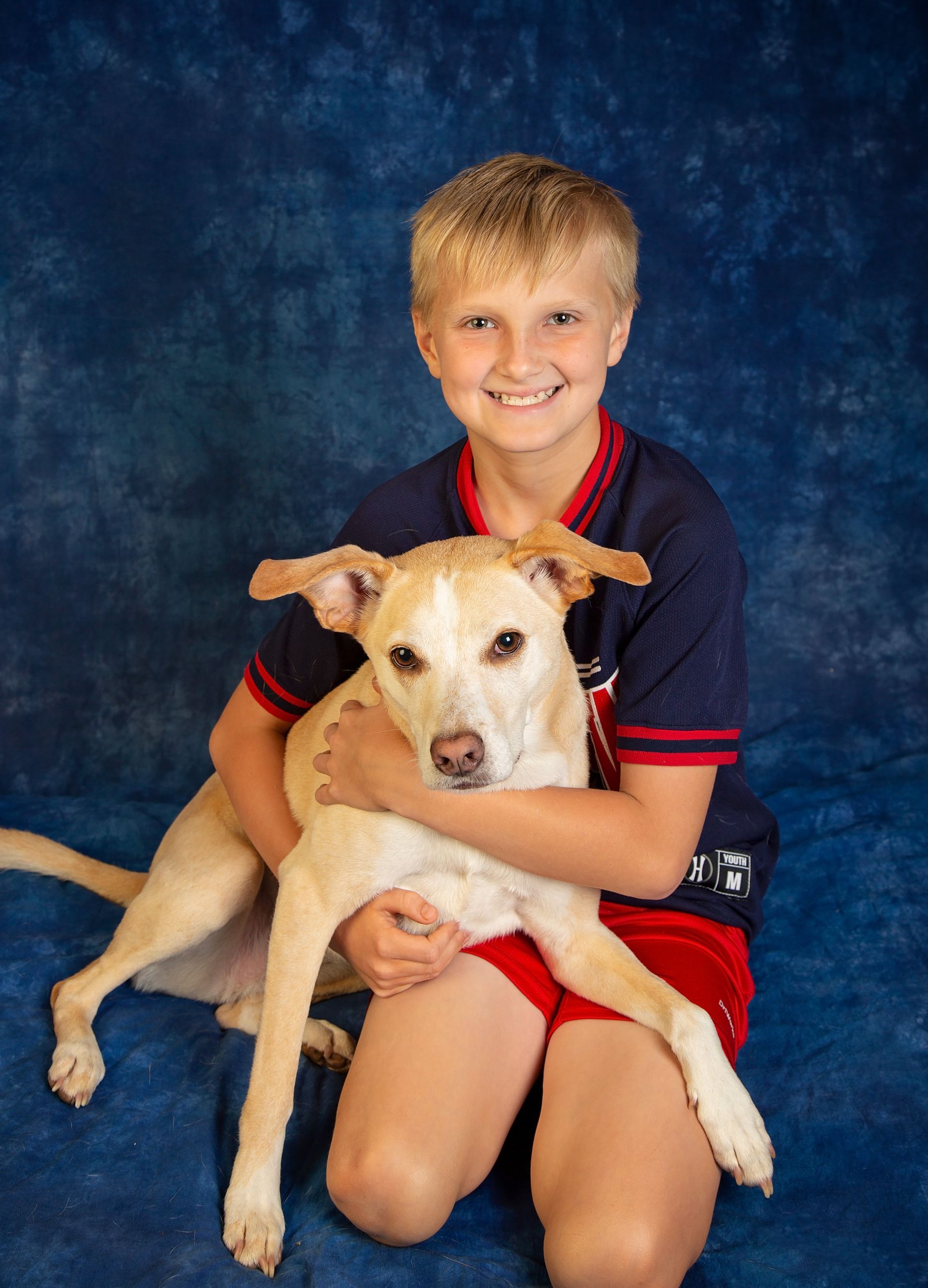 A blond haired boy kneels on the floor holding his dog in his lap.