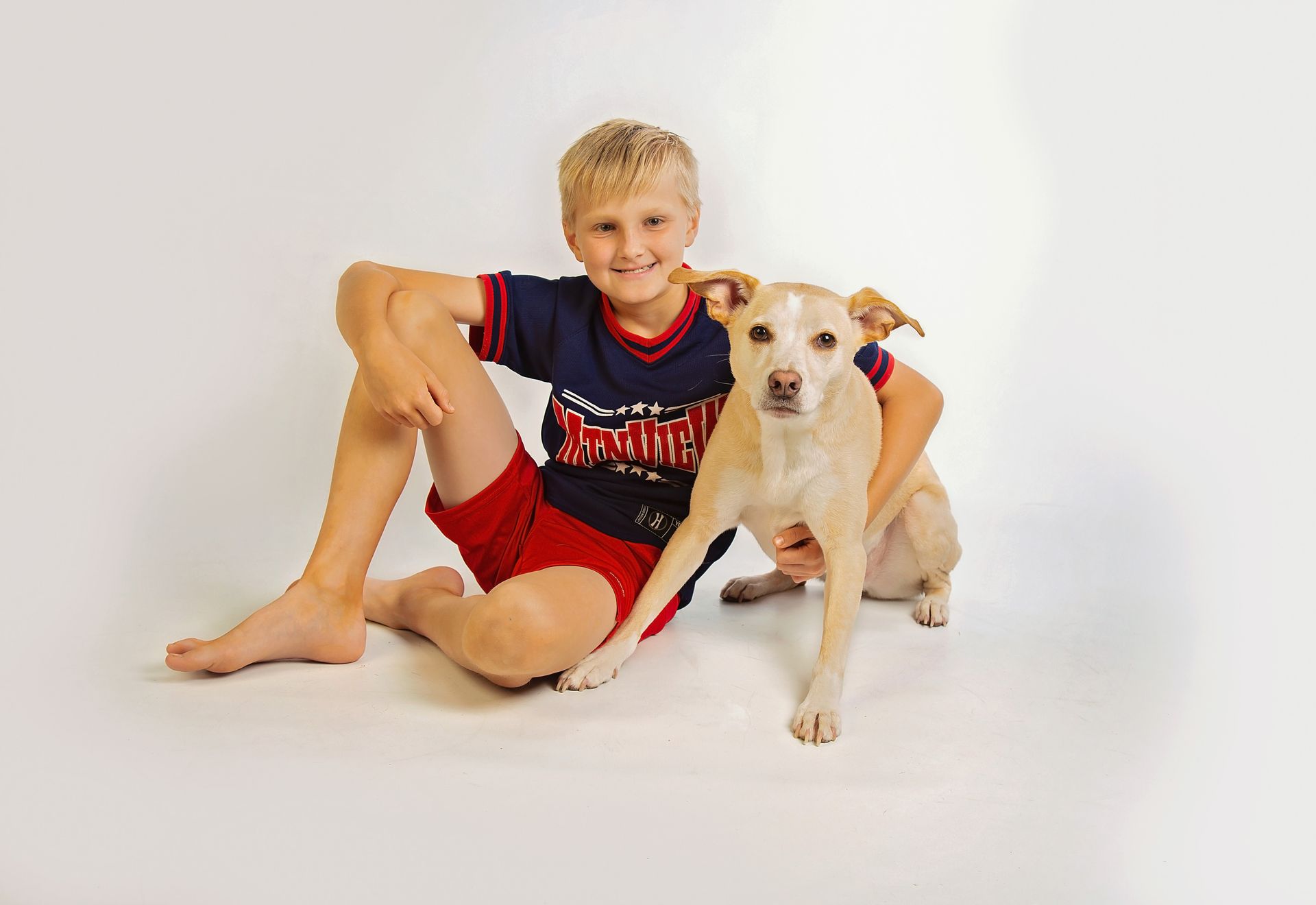 A young blond haired boy sits on the floor with his arm around his dog.