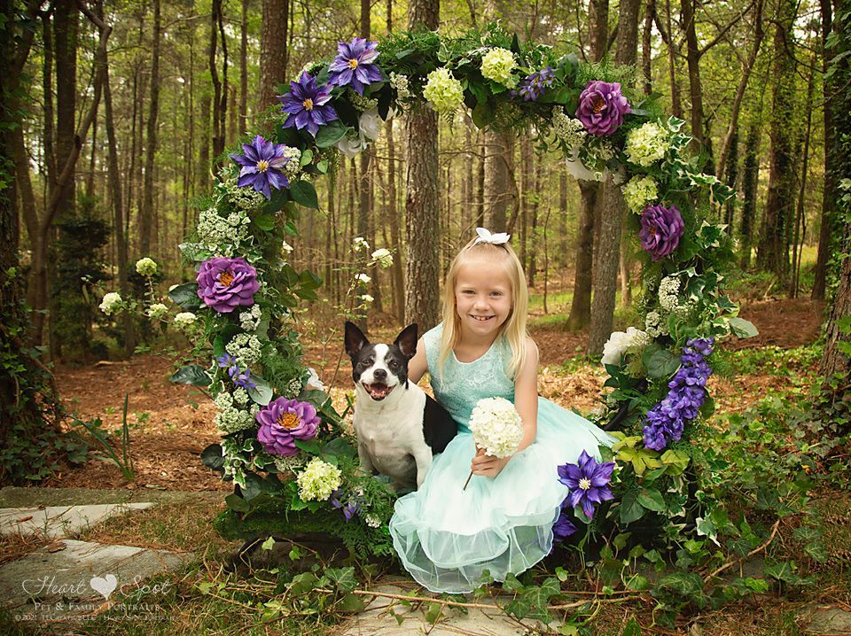 A little girl is sitting in a flower arch with a dog.