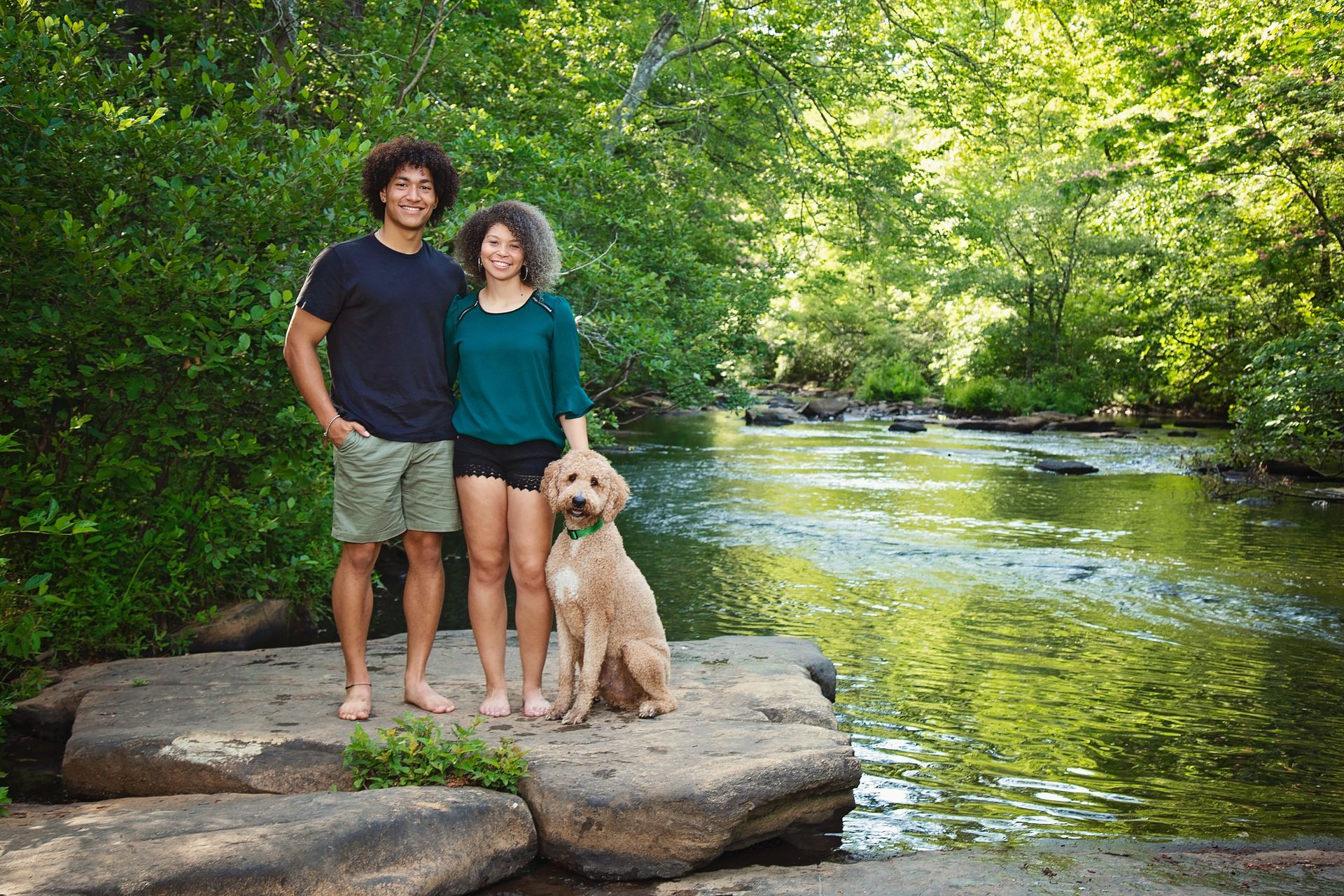 Two teenagers are standing next to a dog on a rock near a river.