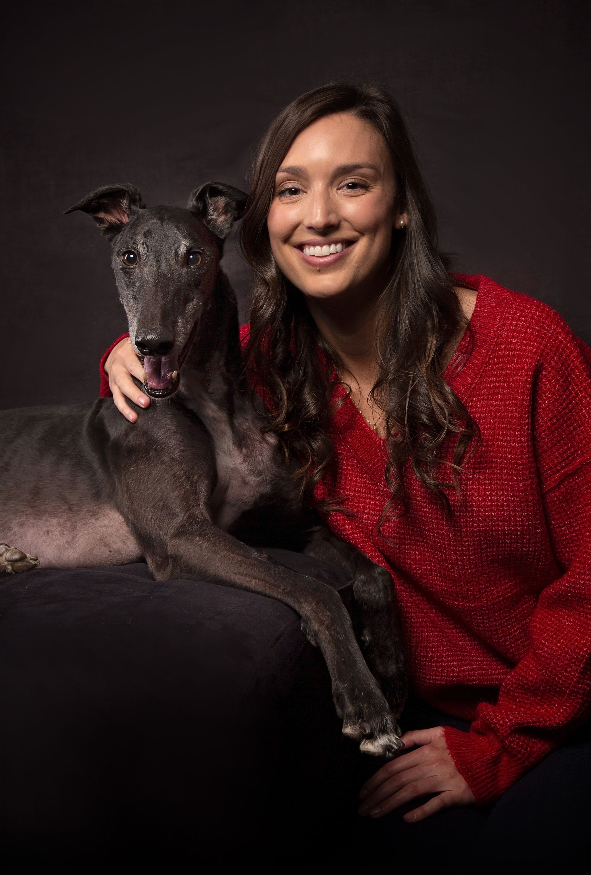 A woman in a red sweater is sitting next to a greyhound dog.