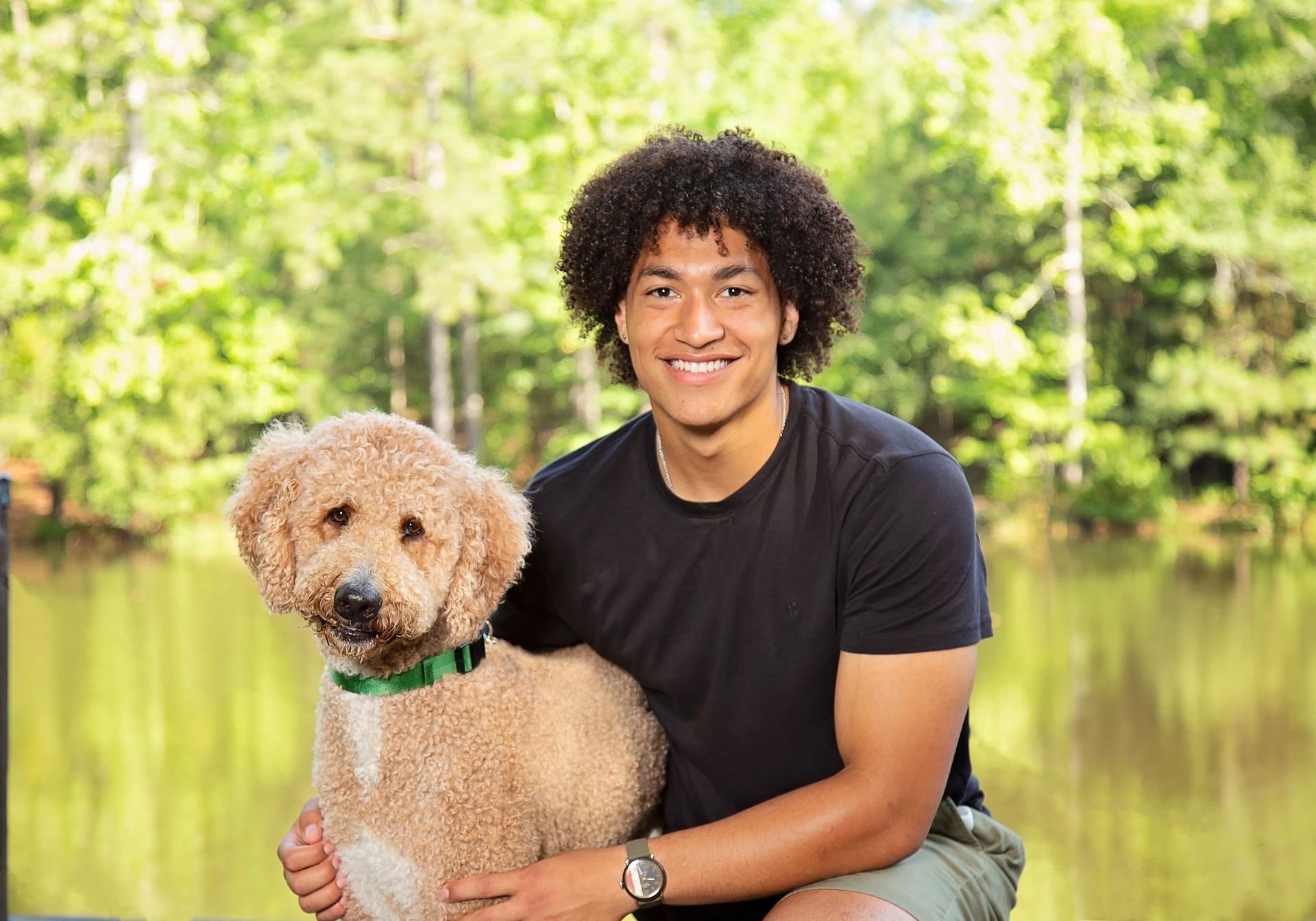 A handsome young man is holding a  brown golden doodle dog in his arms outside on a sunny day.