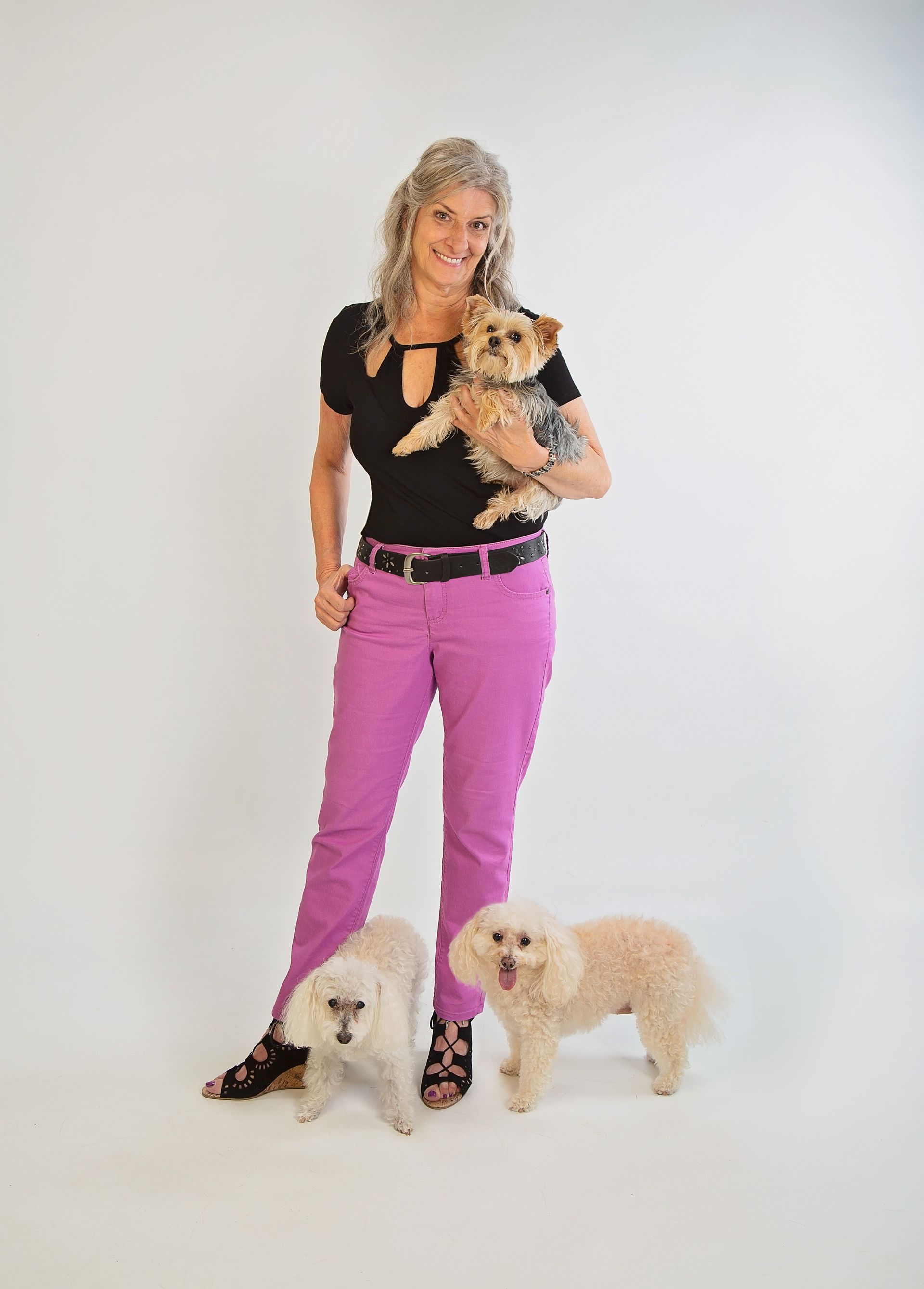 A long blond haired woman in pink pants is standing next to two small dogs while holding a third small dog.