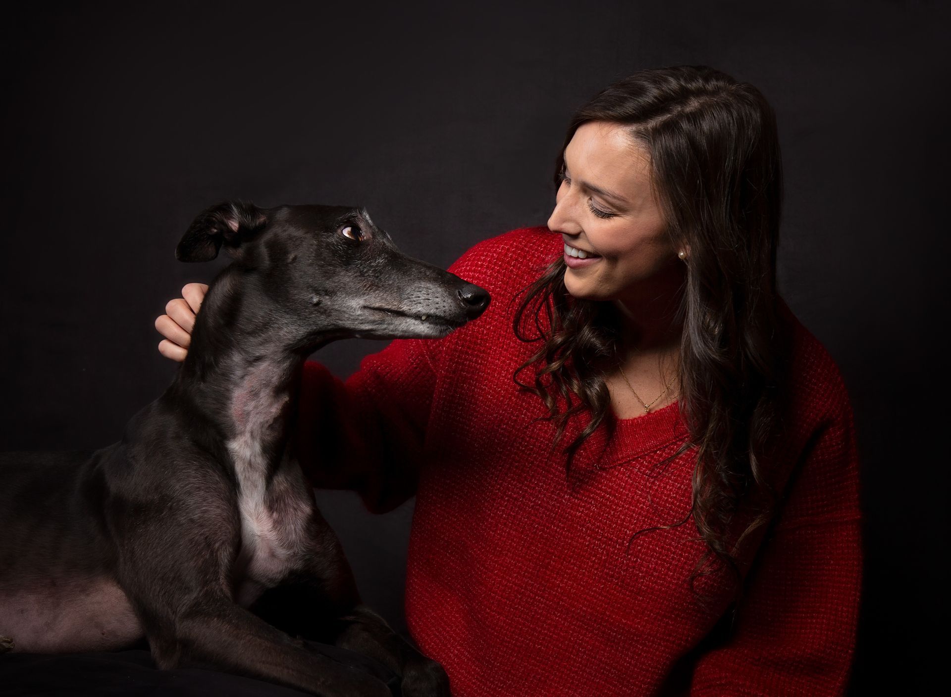 A woman with long brown hair in a red sweater is petting a black greyhound dog while they look into each other's eyes.