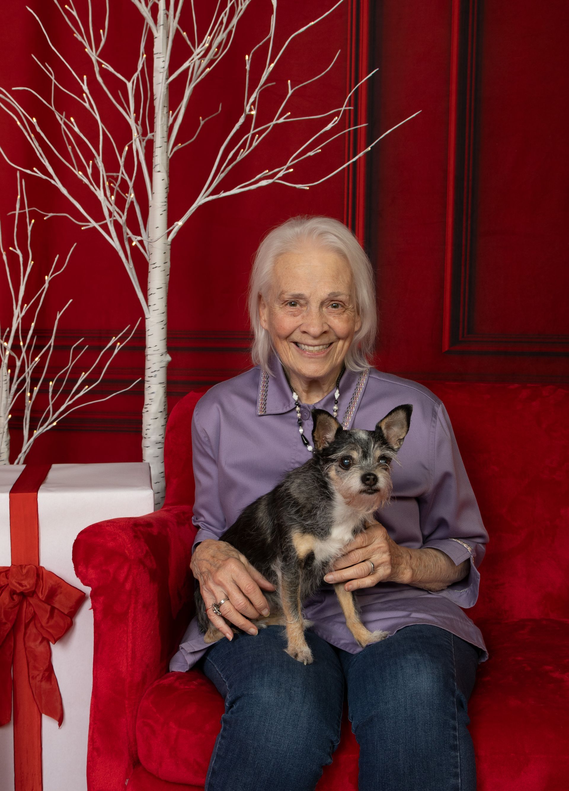 An elderly woman is sitting on a red couch holding a small dog.