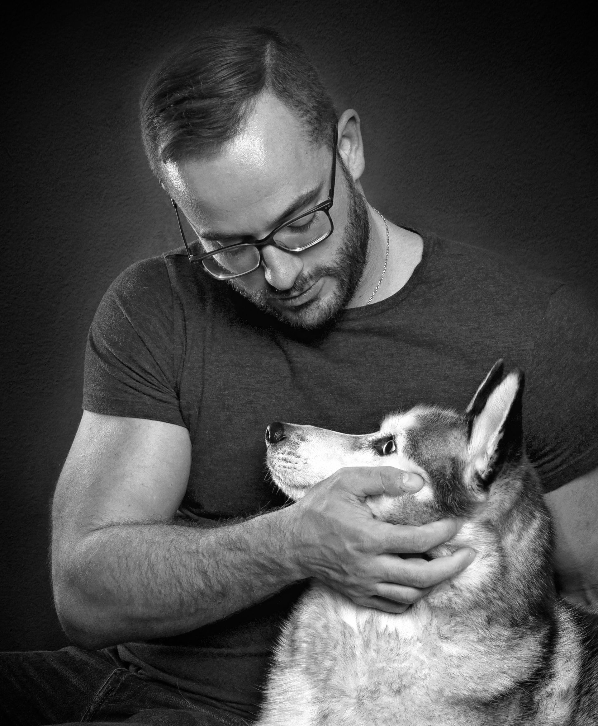 A man with glasses is petting a husky dog in a black and white photo.