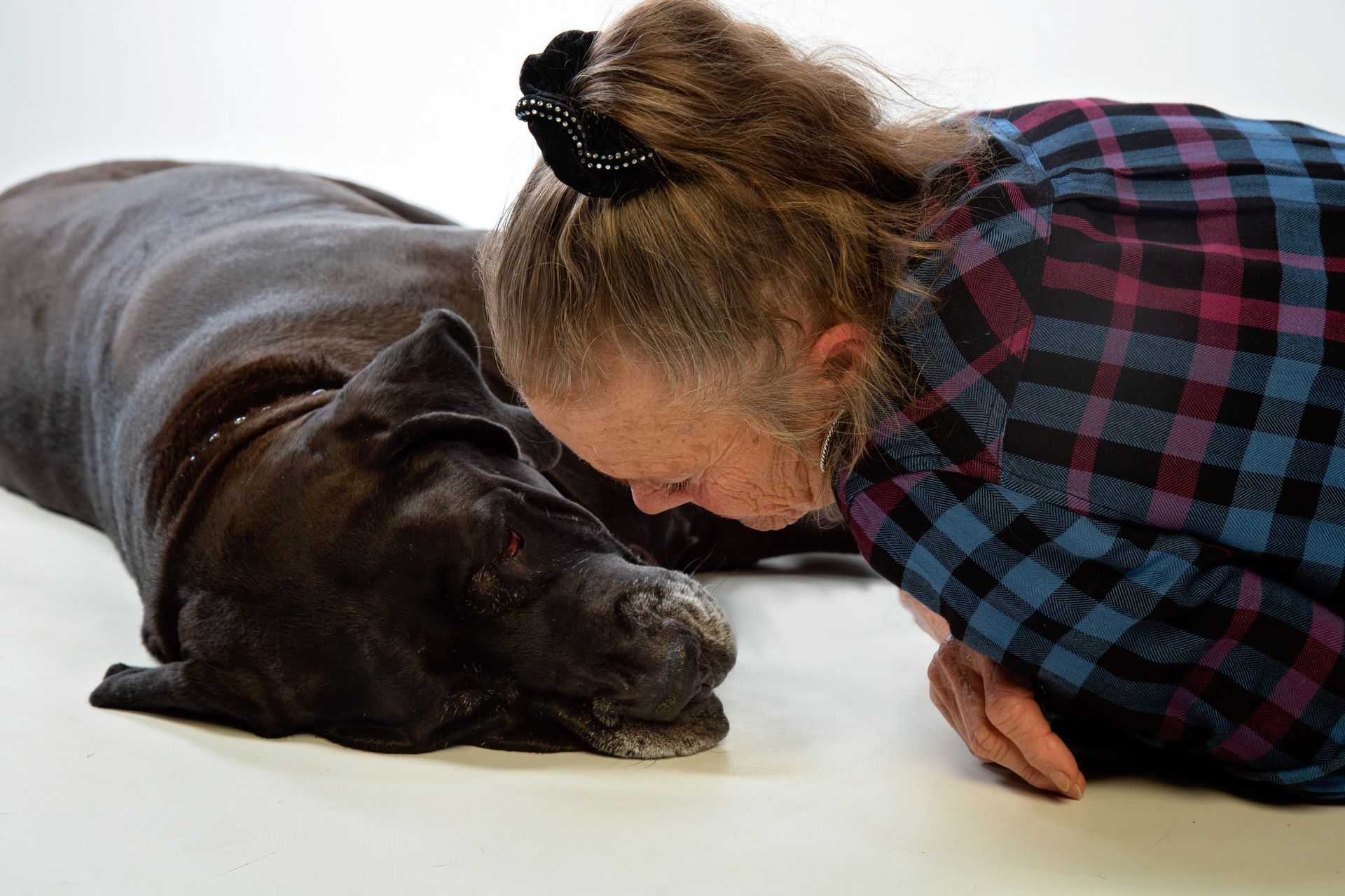 A woman in a plaid shirt is laying down next to a black dog sprawled out on the floor.