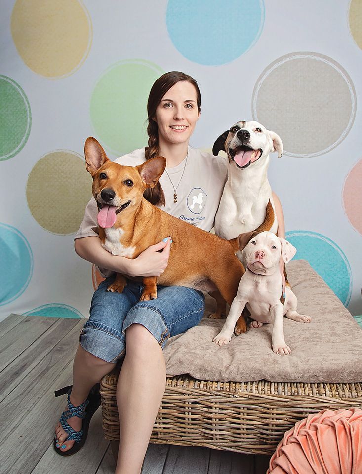 A woman with long braided brown hair is sitting on a wicker chair holding three dogs.