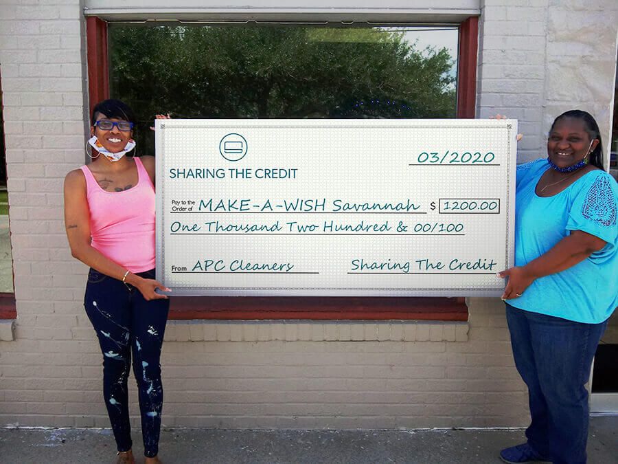 Two women are holding a large check in front of a window.