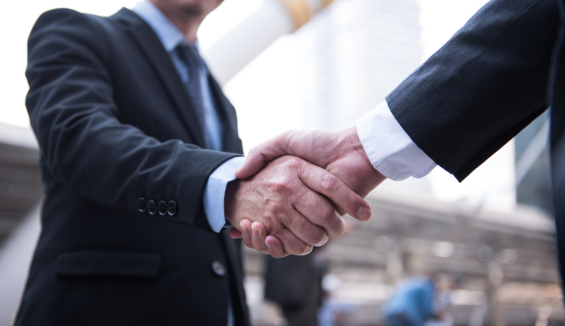 Two men in suits shake hands, business deal. City background, close-up.