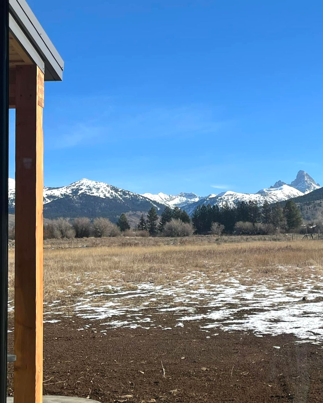 Snowy mountains behind a brown field with a wooden porch post and a clear blue sky.