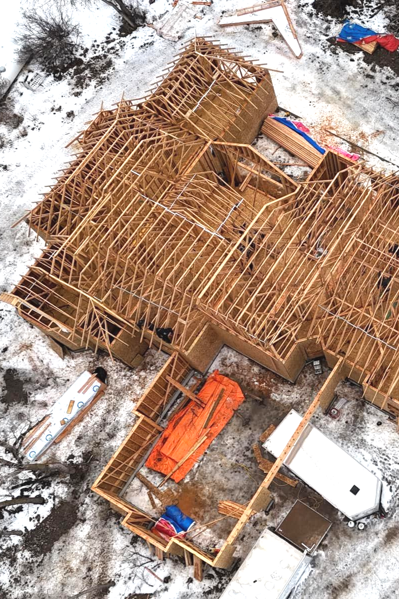Construction site, wooden frames of a house, partially built. Orange tarp and blue materials visible.