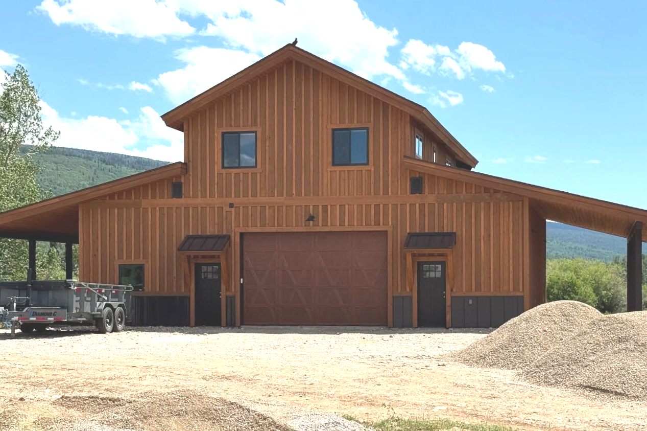 Brown wooden barn with two doors, a large garage door, and blue windows, set in a sunny landscape.