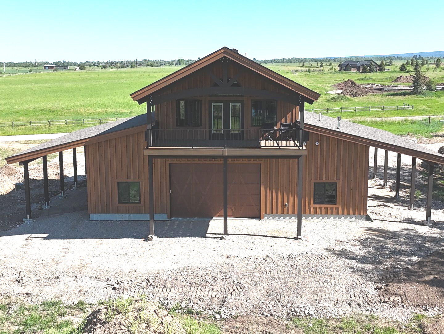Barn-style house with a balcony. Brown exterior with covered side areas, set in a grassy field.