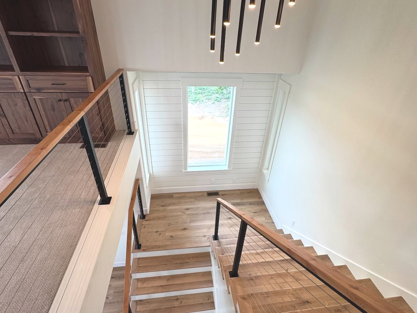 View down a wooden staircase with a black railing, leading to a window.