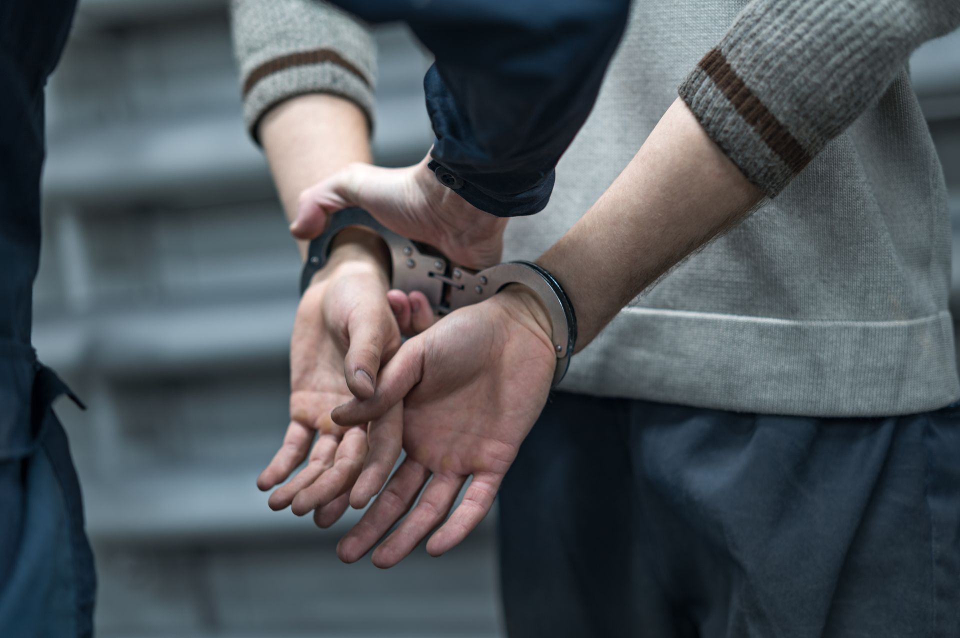 Person in handcuffs with an officer, hands behind back, in front of a gray staircase.