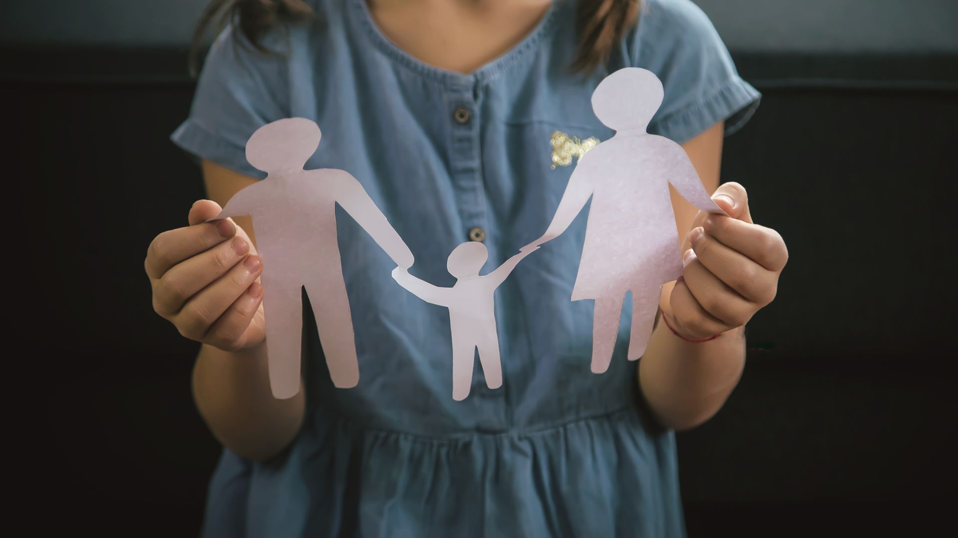 Child holding paper cutouts of a family, against a dark background.