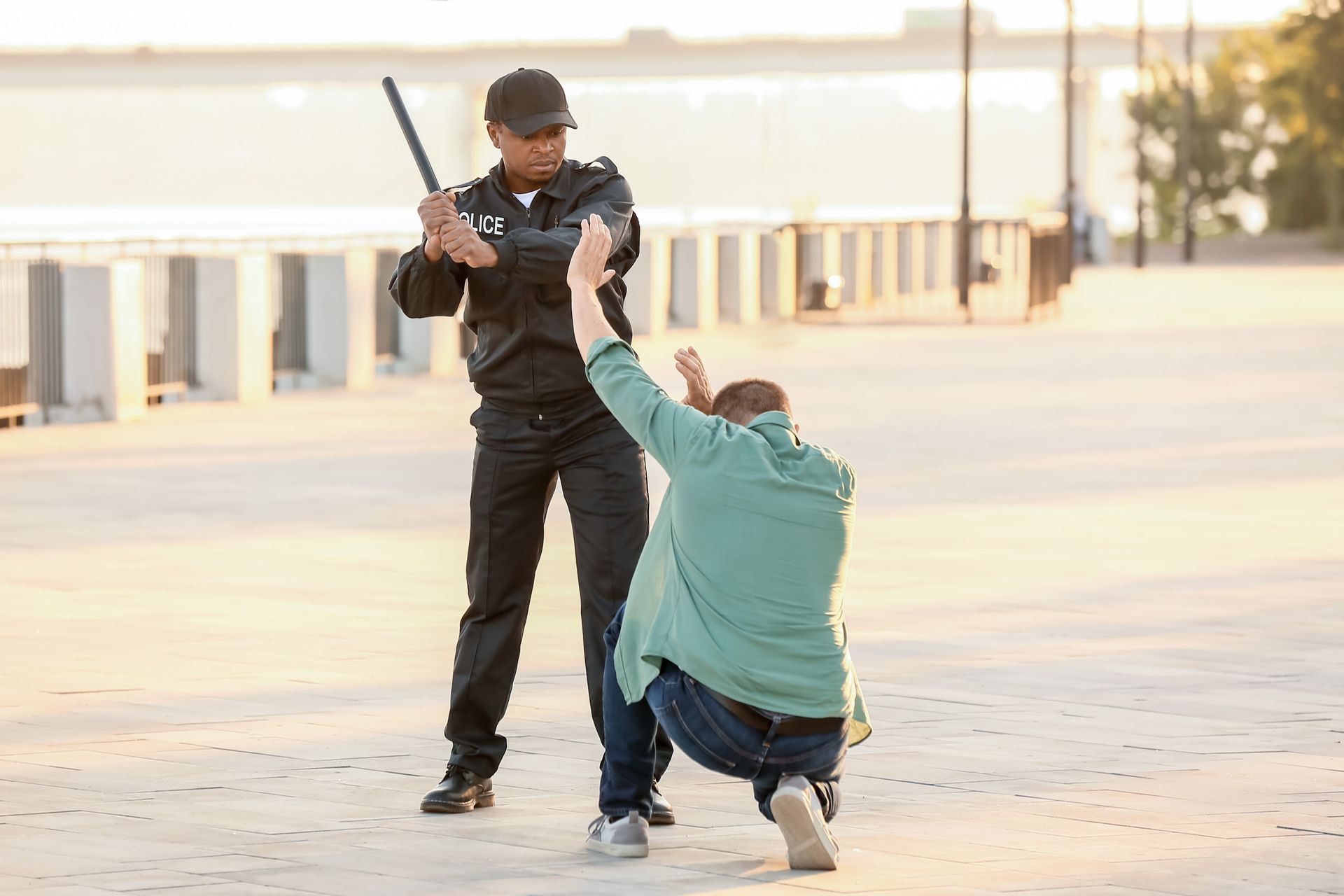 Police officer with baton facing a kneeling person with hands up in a park.