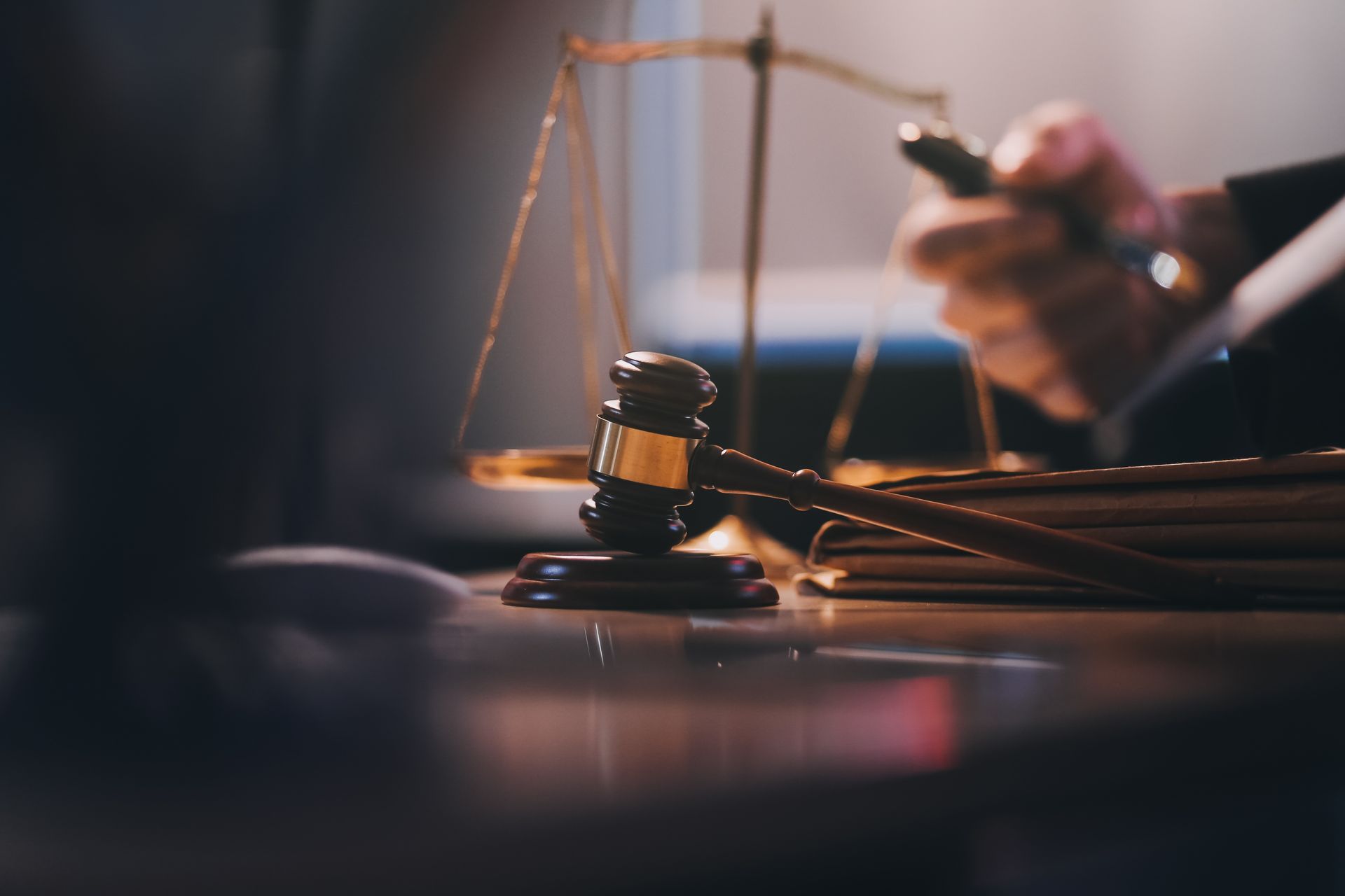 Gavel on a desk next to a balance scale and legal documents; a hand in background.