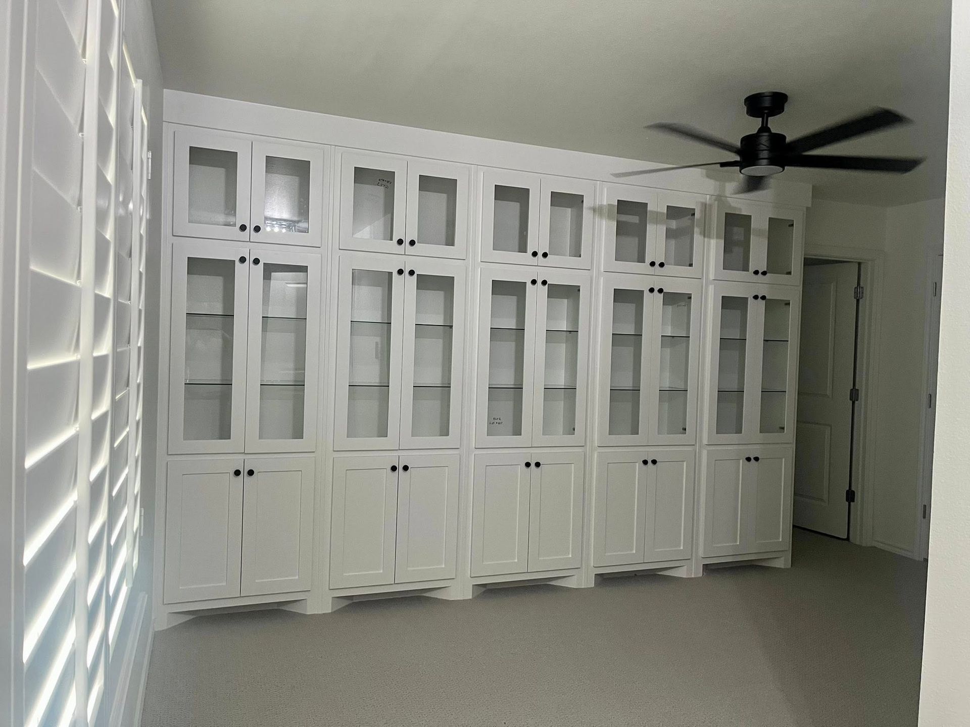 White built-in cabinets with glass doors, a ceiling fan, and carpeted floor in a room.